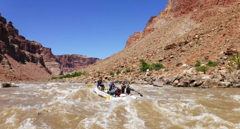 People paddle a raft through whitewater framed by red canyon walls. 